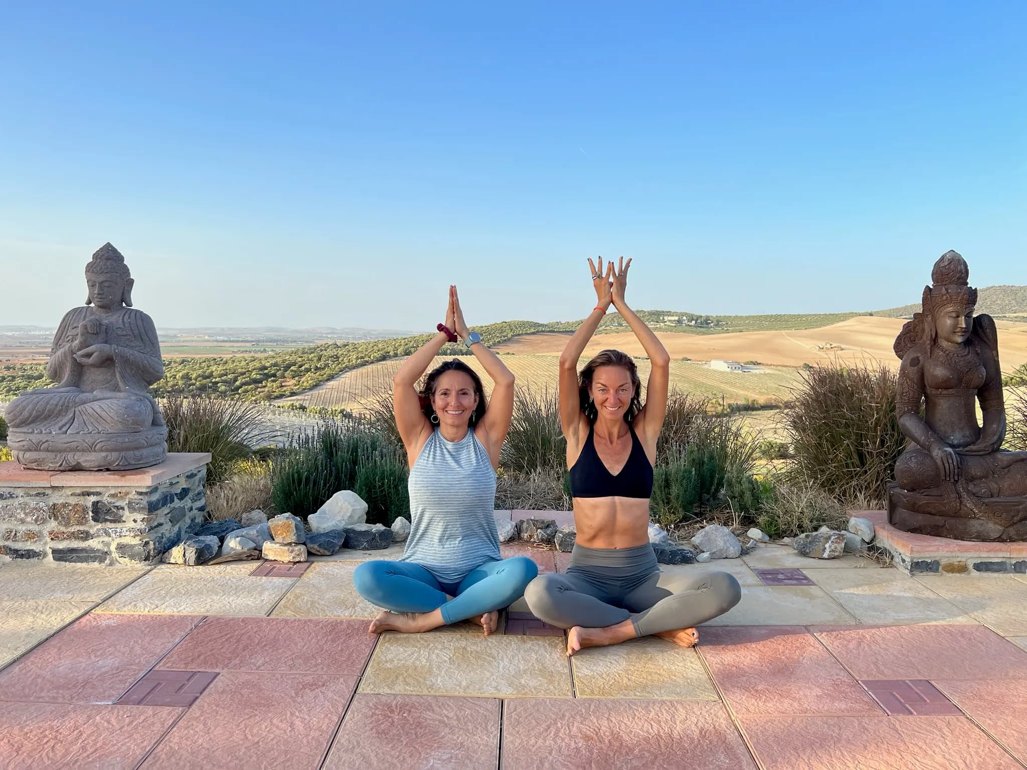 Two people meditating with hands overhead, between Buddhas and hills.